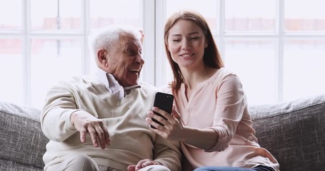 Wall Mural - Happy older 80s grandfather posing for selfie shot on cellphone with smiling attractive grownup granddaughter, resting on sofa. Joyful different generations family having fun, using mobile photo apps.