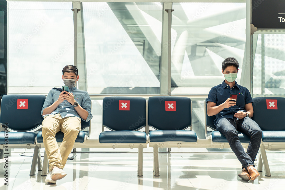 Social distancing, two men wearing face mask sitting keeping distance away from each other to prevent covid19 infection during pandemic. Empty chair seat red cross shows avoidance in airport terminal. - obrazy, fototapety, plakaty 