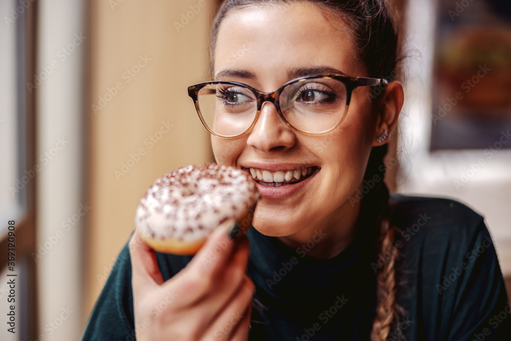 Portrait of smiling teenage girl sitting at pastry shop, holding doughnut and enjoying free time. - obrazy, fototapety, plakaty 