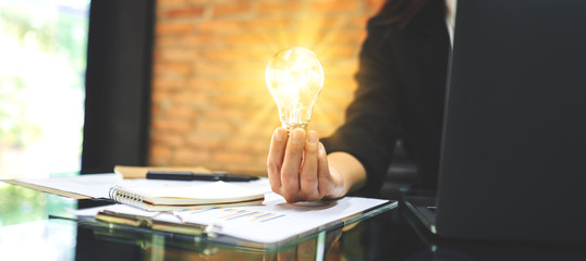 Closeup image of a businesswoman holding a glowing light bulb while working on laptop computer in office