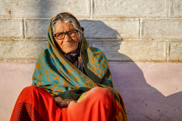 Old aged woman sitting with the support of the wall in the sunset wearing glasses