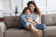 © fizkes - Family portrait smiling mother hugging cute little daughter, sitting on couch at home, looking at camera, happy loving mum holding preschool girl, posing for photo, enjoying tender moment