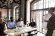 © fizkes - Diverse employees sitting at table, listening to business coach giving flip chart presentation at corporate meeting, confident young mentor team leader training staff, explaining project strategy