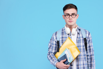 Young man in white t-shirt, checkered shirt, glasses, headphones is holding books, notebooks, standing on blue background. Guy student is ready to study. Education at university concept.