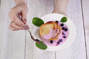 Wall Mural - Closeup girl hand holds a white plate with fritters poured by yogurt with slices of blueberries and mint. A teaspoon in the other hand, a wonderful breakfast and a great start to the day