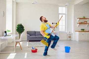 Funny fat man with a beard in an apron dancing cleans the room in the house.