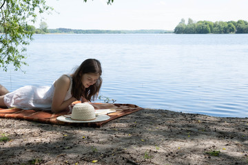 young girl in a white dress lies on the shore of a beautiful lake and reads something