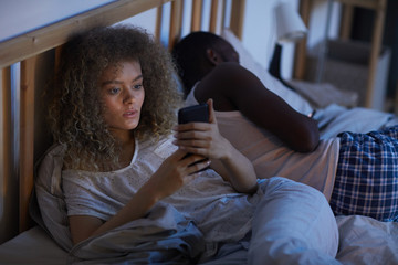 Wall Mural - High angle portrait of young curly-haired woman using smartphone in bed at night while suffering from insomnia, copy space