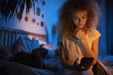 Wall Mural - Portrait of curly-haired young woman sitting on family bed at night and holding smartphone, looking nervous, copy space