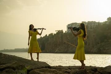 Wall Mural - Two beautiful twin sisters violinists in yellow concert dresses are playing electric violins on sunset on rocks by the Mediterranean sea in old town of Antalya Kaleici Turkey.