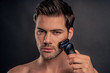 © Roman - Handsome young bearded man isolated. Portrait of topless muscular man is standing on gray background with electric Shaver in hand while shaving. Men care concept.