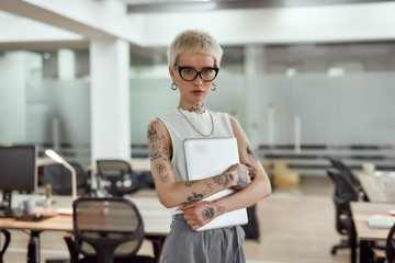 Digital technologies. Young, stylish and attractive tattooed business woman holding her laptop and looking at camera while standing in the modern coworking space