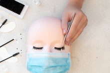 A Beauty Mannequin Training Head Wearing A Protective Mask With A Person Practising Lash Extension Techniques.  Beauty Treatment Tools Arranged On Left Side Of Background Frame. 