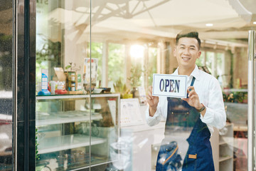 Poster - Portrait of cheerful young Vietnamese waiter hanging open sign on glass wall of cafe