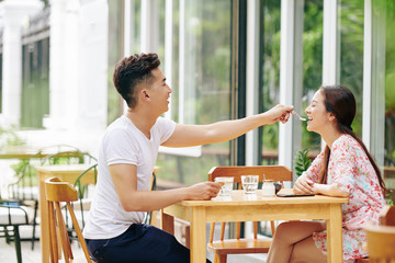 Wall Mural - Happy smiling young Vietnamese man feeding his girlfriend with spoon of yogurt