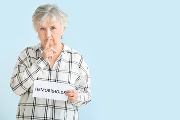 Poster - Elderly woman holding paper with text HEMORRHOIDS and showing silence gesture on color background