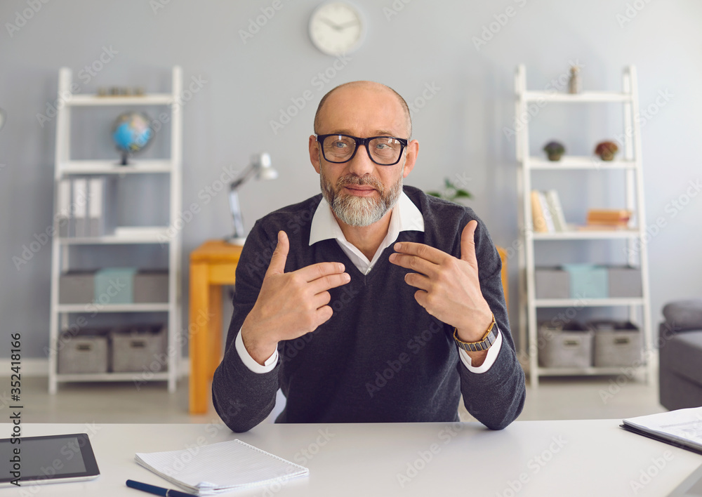 Mature man with a gray beard looks at the camera, speaks with his partner sitting at a table in the office at home. - obrazy, fototapety, plakaty 