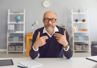 Mature man with a gray beard looks at the camera, speaks with his partner sitting at a table in the office at home.