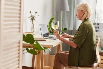 Side view of mature woman sitting at the table with wineglass of wine and using laptop at home