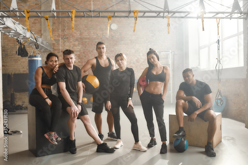 Full length shot of a group of sportive people in black sportswear looking at camera while standing at industrial gym. Group training, teamwork concept