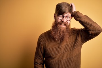 Poster - Handsome Irish redhead man with beard wearing glasses and winter sweater over yellow background confuse and wonder about question. Uncertain with doubt, thinking with hand on head. Pensive concept.