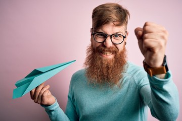 Sticker - Irish redhead man with beard holding paper plane over pink isolated background annoyed and frustrated shouting with anger, crazy and yelling with raised hand, anger concept