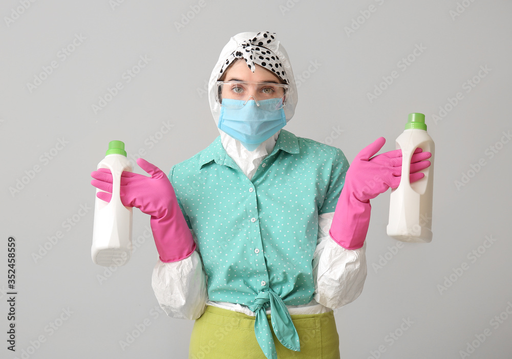 Housewife in protective costume and with detergents on grey background