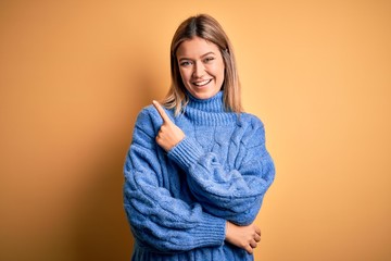 Poster - Young beautiful blonde woman wearing turtleneck sweater over yellow isolated background cheerful with a smile of face pointing with hand and finger up to the side with happy and natural expression