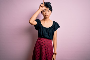 Wall Mural - Young beautiful asian girl wearing casual dress standing over isolated pink background making fun of people with fingers on forehead doing loser gesture mocking and insulting.