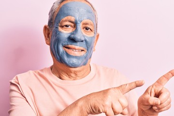 Poster - Senior man with grey hair wearing mud mask smiling and looking at the camera pointing with two hands and fingers to the side.