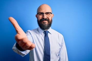 Poster - Handsome business bald man with beard wearing elegant tie and glasses over blue background smiling friendly offering handshake as greeting and welcoming. Successful business.