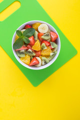 Wall Mural - Fruit salad in the white  bowl on the green cutting board on the yellow background. Top view. Copy space. Location vertical.