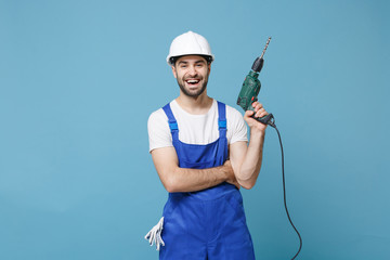 Cheerful young man in coveralls protective helmet hardhat hold electric drill isolated on pastel blue background in studio. Instruments accessories for renovation apartment room. Repair home concept.