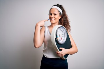 Wall Mural - Young beautiful woman with curly hair holding weighing machine over white background smiling cheerful showing and pointing with fingers teeth and mouth. Dental health concept.