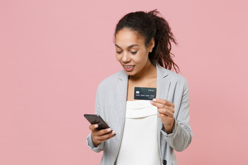 Wall Mural - Smiling african american business woman in grey suit, white shirt isolated on pink background. Achievement career wealth business concept. Mock up copy space. Using mobile phone hold credit bank card.