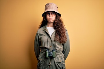 Poster - Young beautiful tourist woman on vacation wearing explorer hat and binoculars puffing cheeks with funny face. Mouth inflated with air, crazy expression.