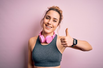 Young beautiful blonde sportswoman wearing sportswear listening to music using headphones doing happy thumbs up gesture with hand. Approving expression looking at the camera showing success.