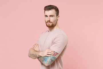 Handsome young bearded tattooed man guy in pastel casual t-shirt posing isolated on pink wall background studio portrait. People emotions lifestyle concept. Mock up copy space. Holding hands crossed.