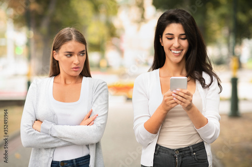 Cuadros en Lienzo Jealous Girl Looking At Friend's Phone Walking Together Outdoors