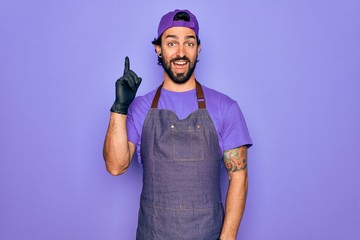 Young handsome hispanic man with bear wearing professional apron working as tattoo artist pointing finger up with successful idea. Exited and happy. Number one.