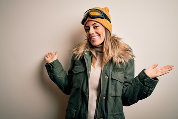 Poster - Young brunette skier woman wearing snow clothes and ski goggles over white background smiling showing both hands open palms, presenting and advertising comparison and balance