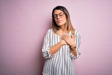 Sticker - Young beautiful woman wearing casual striped t-shirt and glasses over pink background smiling with hands on chest with closed eyes and grateful gesture on face. Health concept.