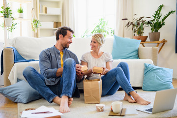 Wall Mural - Happy couple sitting on floor indoors at home, eating hamburgers.