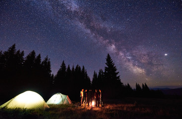 Wall Mural - group of tourists are standing around burning fire near the illuminated tents against backdrop pine 