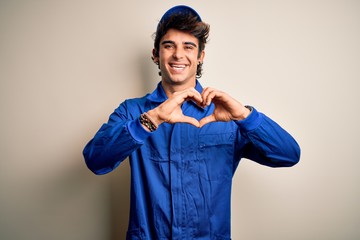 Wall Mural - Young mechanic man wearing blue cap and uniform standing over isolated white background smiling in love doing heart symbol shape with hands. Romantic concept.