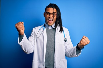 Wall Mural - Young african american doctor man with dreadlocks wearing stethoscope and glasses celebrating surprised and amazed for success with arms raised and open eyes. Winner concept.