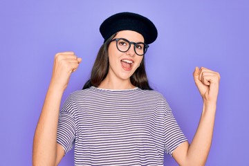 Poster - Young beautiful brunette woman wearing glasses and french beret over purple background celebrating surprised and amazed for success with arms raised and open eyes. Winner concept.