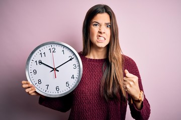 Sticker - Young beautiful girl doing countdown holding big clock over isolated pink background annoyed and frustrated shouting with anger, crazy and yelling with raised hand, anger concept