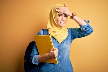 Poster - Young student girl wearing muslim hijab and backpack holding book over yellow background stressed with hand on head, shocked with shame and surprise face, angry and frustrated. Fear and upset