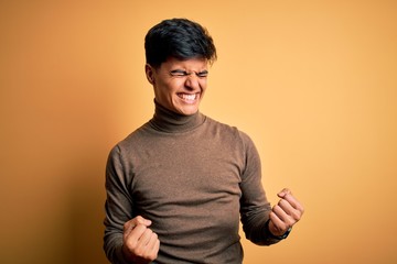 Poster - Young handsome man wearing casual turtleneck sweater over isolated yellow background very happy and excited doing winner gesture with arms raised, smiling and screaming for success. Celebration